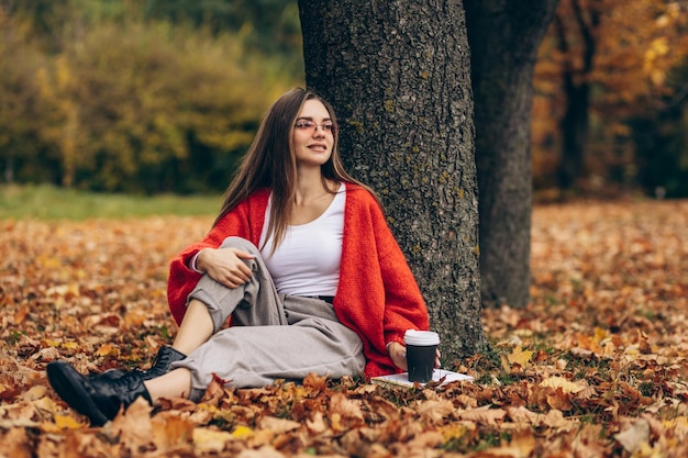 Vrouw zit onder de boom op gras bedekt met herfstbladeren en drinkt koffie