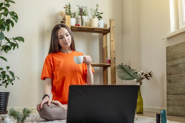 Vrouw zit op de grond voor een laptop in de lotushouding met een kopje thee in haar handen en kijkt naar het scherm Concept van afstands- of videoleren van meditatieyoga