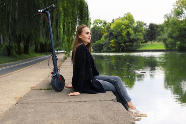 Vrouw zit op de rivieroever naast een elektrische scooter Een wandeling door de stad op een vrije dag Elektrisch vervoer