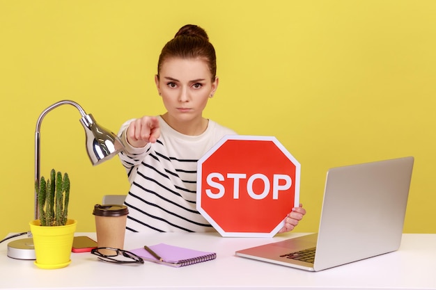 Vrouw zit op de werkplek voor een laptop met een rood stopbord en wijst met de vinger naar de camera