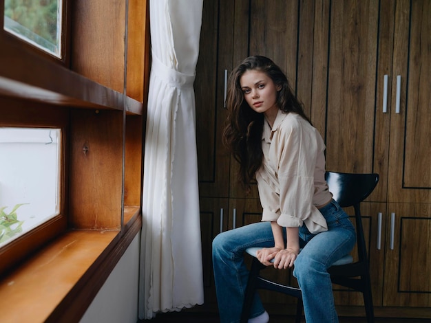 Vrouw zit op een stoel in het interieur van het huis bij het raam comfort en gezelligheid levensstijl ontspanning op het weekend bedachtzame uitstraling van een mooi meisje