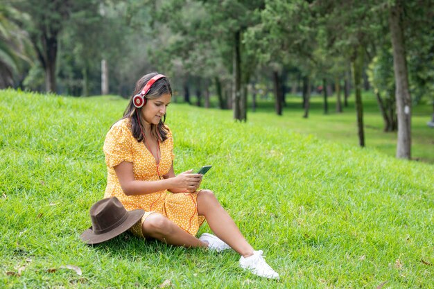 Vrouw zit op het gras en luistert naar muziek met een koptelefoon terwijl ze sms't met haar mobiele telefoon