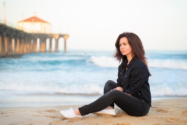 Vrouw zit op het zand voor een pier op Manhattan Beach in Los Angeles in de vroege ochtend