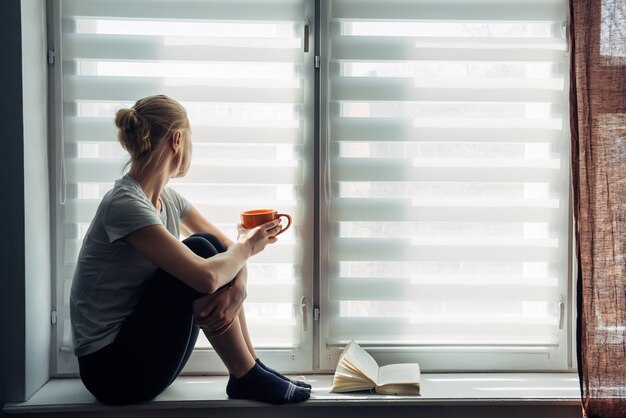 Vrouw zit op vensterbank, leest een boek, drinkt thee