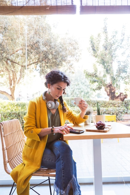 Vrouw zit voor het raam van een cafetaria en kijkt naar haar mobiel