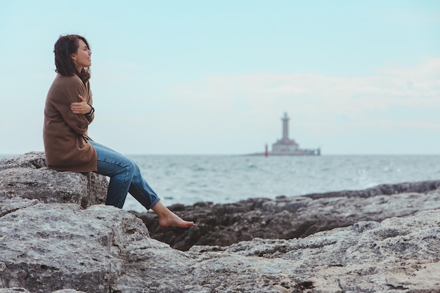 Vrouw zitten door rotsachtige zee strand in natte jeans vuurtoren op background