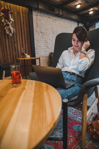 Vrouw zittend in grote stoel in café met laptop luisteren muziek in headset fruit thee drinken