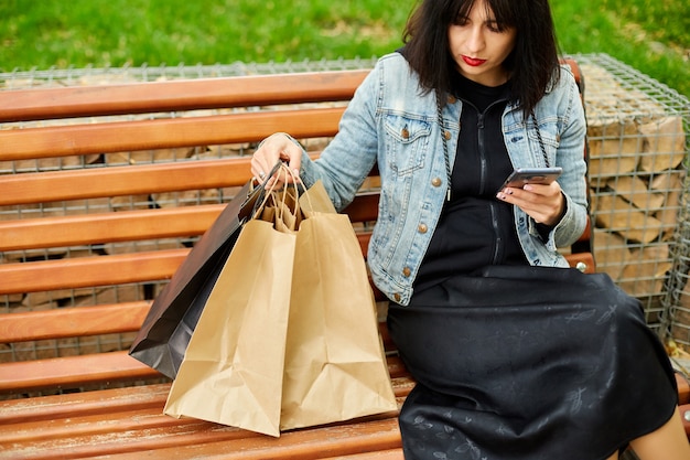 Foto vrouw zittend op bankje park boodschappentassen, smartphone in handen houden, sms-bericht typen