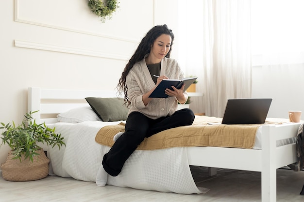 Vrouw zittend op bed met behulp van computer en schrijven in notitieblok