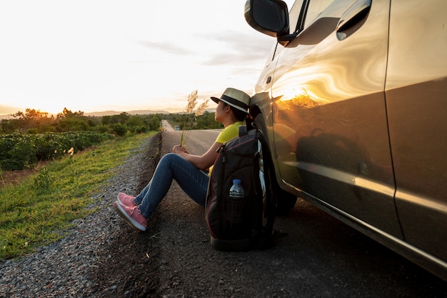 Vrouw zittend op de weg bij zonsondergang met auto's en rugzakken Concept van vakantie en reizen