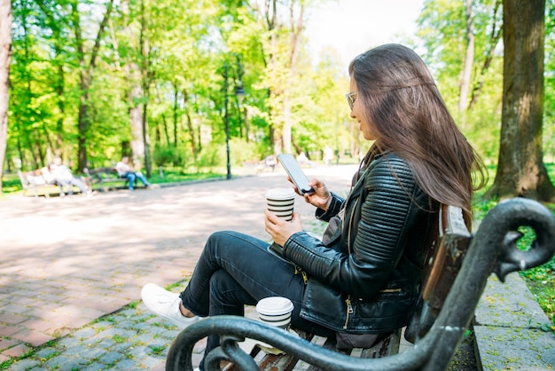 Vrouw zittend op een bankje in het stadspark koffie drinken op internet surfen via mobiel