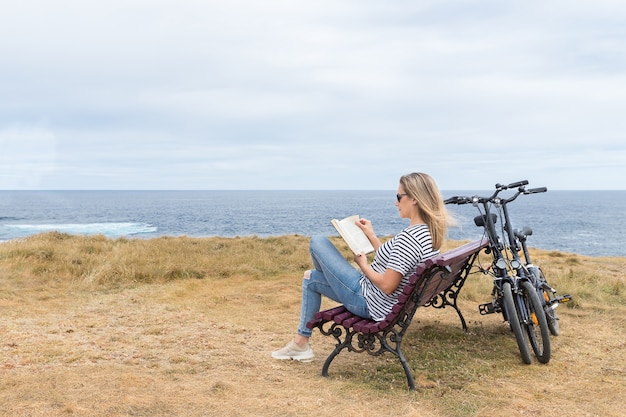 Vrouw zittend op een bankje voor de zee een boek lezen en twee fietsen tegen de bank geleund