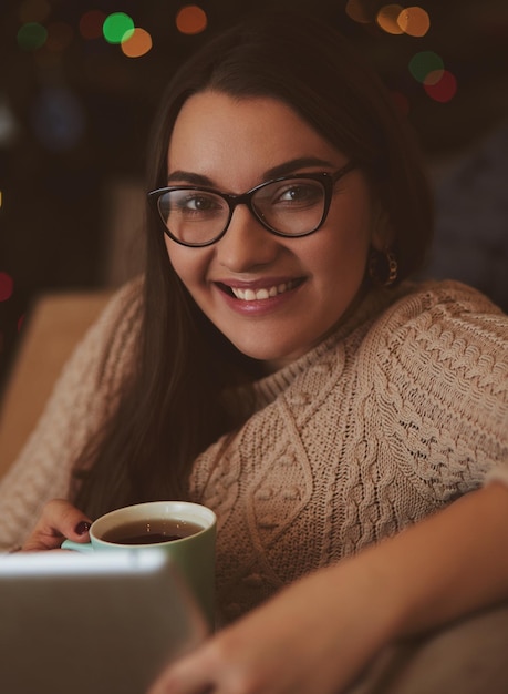 Vrouw zittend op het tapijt in de buurt van de bank met behulp van een tablet, koffie drinken uit een kopje. Online onderwijsconcept.