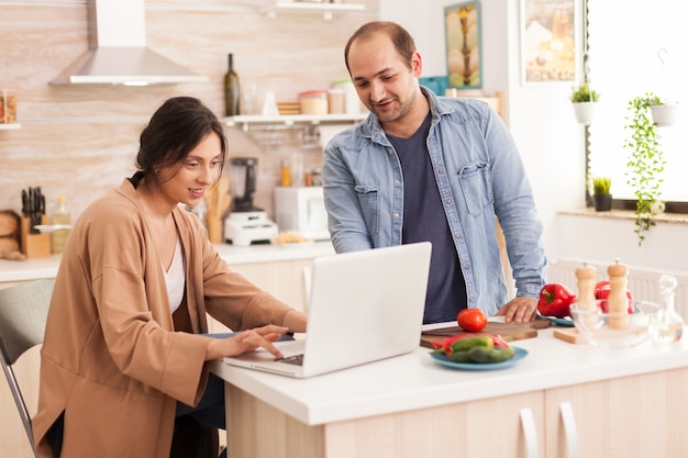 Vrouw zoekt salade recept op laptop in keuken samen met man. man helpt vrouw om gezond biologisch diner te bereiden, samen te koken. romantische vrolijke liefdesrelatie