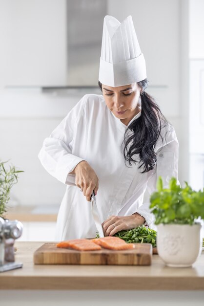 Vrouwelijk chef-kokberoep aan het werk als vrouw in uniform en witte hoed snijdt zalm op een houten plank in een professionele keuken