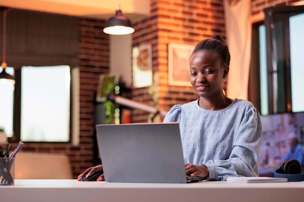 Vrouwelijke Afro-Amerikaanse student op afstand typen op laptop in kamer met modern interieur. Glimlachende freelance copywriter met behulp van draagbare computer in thuiskantoor met prachtig warm zonsonderganglicht