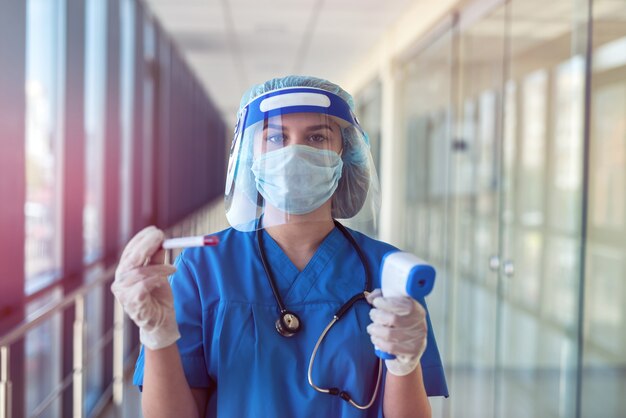 Vrouwelijke arts die een beschermend masker en gezichtsschild draagt met behulp van een infraroodthermometer om de lichaamstemperatuur in de kliniek te controleren. covid19