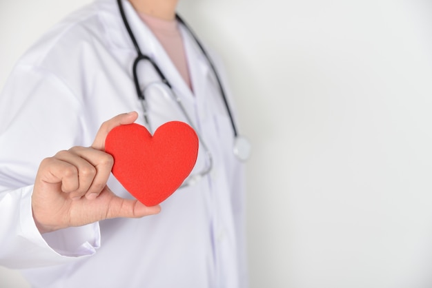 Vrouwelijke arts die met stethoscoop rood hart in haar hand op witte achtergrond houdt. Medische en gezondheidsvoorwaarden.