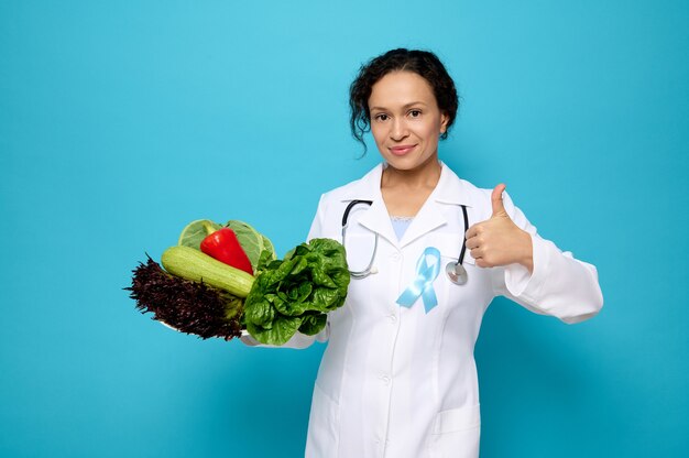 Vrouwelijke arts in witte medische jurk, gekleed in blauw satijnen lint, symbolische strikkleur die het bewustzijn op diabetesdag verhoogt, een bord met rauwe veganistische gezonde maaltijd vasthoudt en duim omhoog kijkt naar de camera