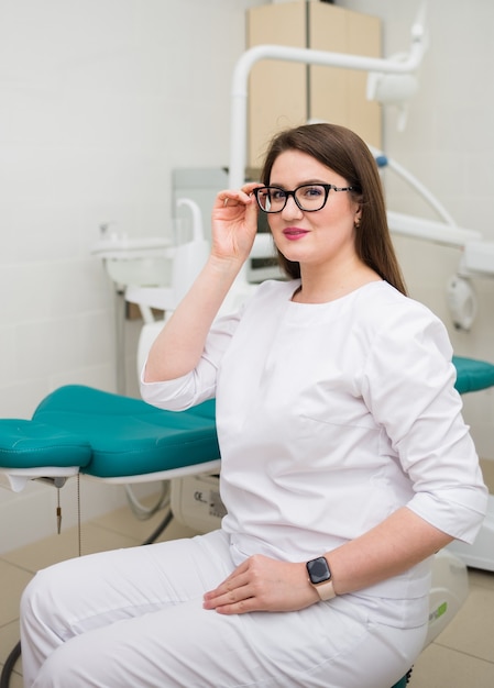 Vrouwelijke arts met een bril en een wit medisch pak zit in de buurt van een tandartsstoel in de kliniek