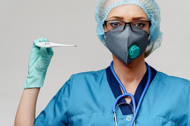 Vrouwelijke arts met stethoscoop die beschermend masker en latexhandschoenen over lichtgrijze achtergrondholdingsthermometer dragen