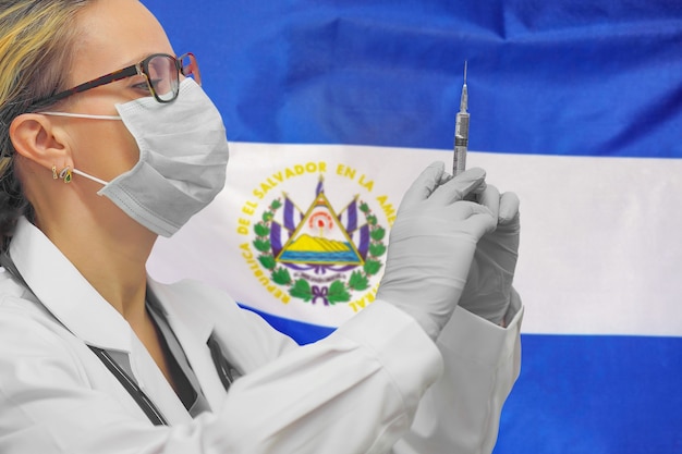 Vrouwelijke arts of verpleegster die in handschoenen spuit voor vaccinatie houdt tegen de achtergrond van de vlag van El Salvador