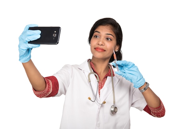 Vrouwelijke arts selfie met stethoscoop en injectie door smartphone op witte achtergrond.