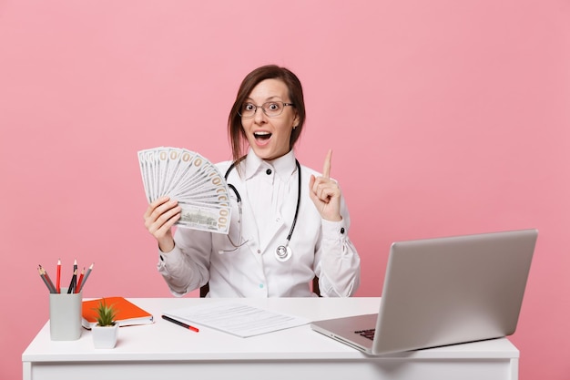 Vrouwelijke arts zit aan bureau werk op computer met medisch document contant geld in ziekenhuis geïsoleerd op pastel roze muur achtergrond. Vrouw in medische toga glazen stethoscoop. Gezondheidszorg geneeskunde concept