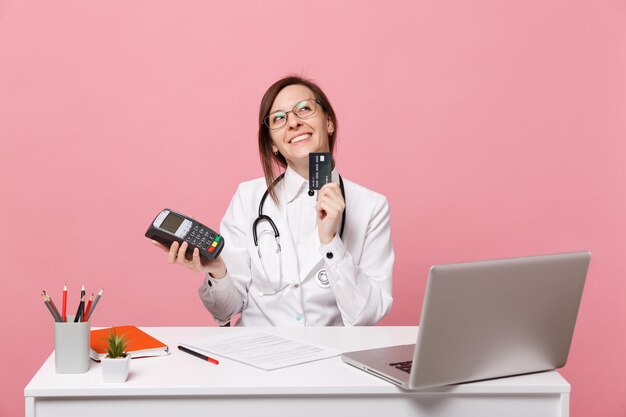 Vrouwelijke arts zit aan bureau werk op computer met medisch document creditcard in ziekenhuis geïsoleerd op pastel roze muur achtergrond. vrouw in medische toga glazen stethoscoop. gezondheidszorg geneeskunde concept