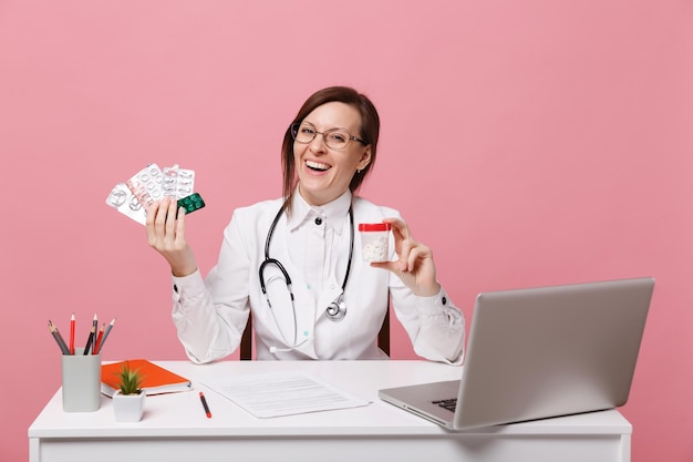Vrouwelijke arts zit aan bureau werk op computer met medisch document houd pillen in ziekenhuis geïsoleerd op pastel roze muur achtergrond. Vrouw in medische toga glazen stethoscoop. Gezondheidszorg geneeskunde concept.