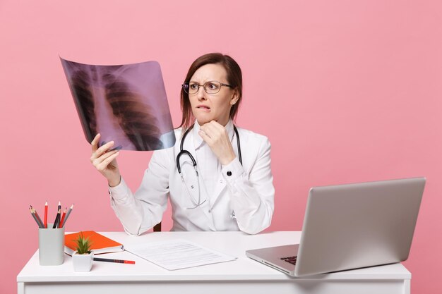 Vrouwelijke arts zit aan bureau werk op computer met medisch document houdt röntgenfoto in ziekenhuis geïsoleerd op pastel roze muur achtergrond. Vrouw in medische toga glazen stethoscoop. Gezondheidszorg geneeskunde concept.