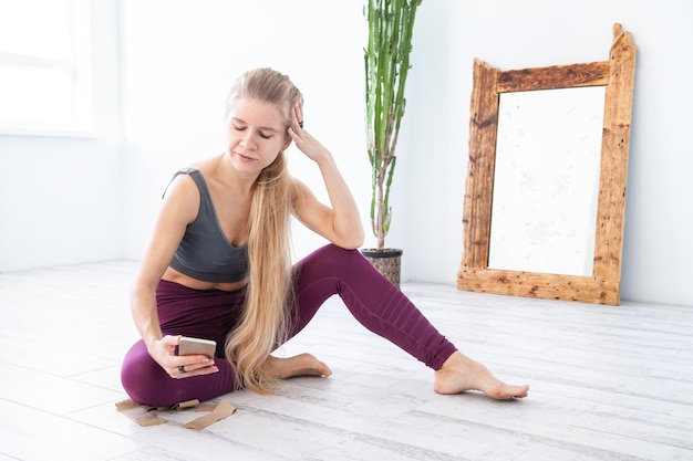 Foto vrouwelijke atleet op blote voeten in sportkleding die op de vloer rust en smartphone doorbladert tijdens de pauze in de training thuis