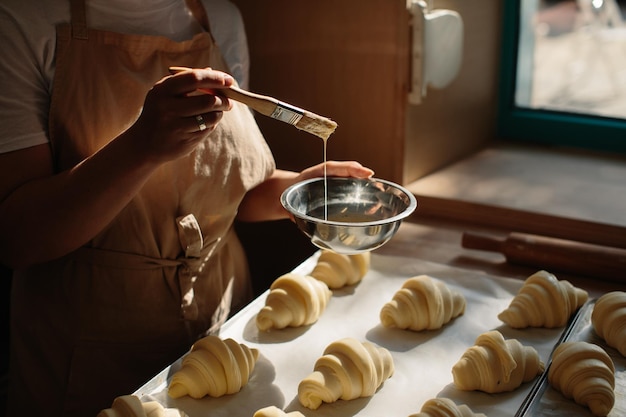 Vrouwelijke bakker smeert rauwe croissants met een borstel in de dooier het kookproces