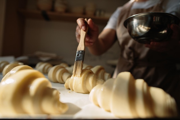 Vrouwelijke bakker smeert rauwe croissants met een borstel in de dooier het kookproces