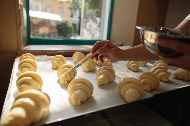 Vrouwelijke bakker smeert rauwe croissants met een borstel in de dooier het kookproces