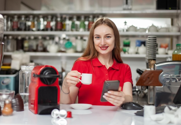 Vrouwelijke barista drinkt koffie en gebruikt smartphone op het werk