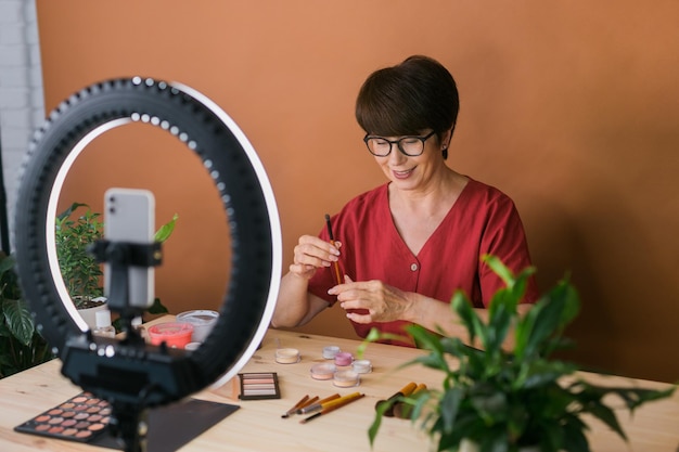 Vrouwelijke beautyblogger of influencer van middelbare leeftijd met make-upproduct dat video opneemt in de kamer thuis Ringlamp en smartphone gebruiken
