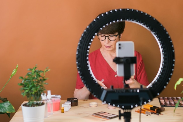 Foto vrouwelijke beautyblogger of influencer van middelbare leeftijd met make-upproduct dat video opneemt in de kamer thuis ringlamp en smartphone gebruiken