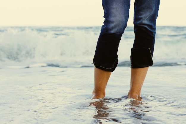 Vrouwelijke benen in jeans die zich in het zeewater op de achtergrond van een brekende golf bevinden