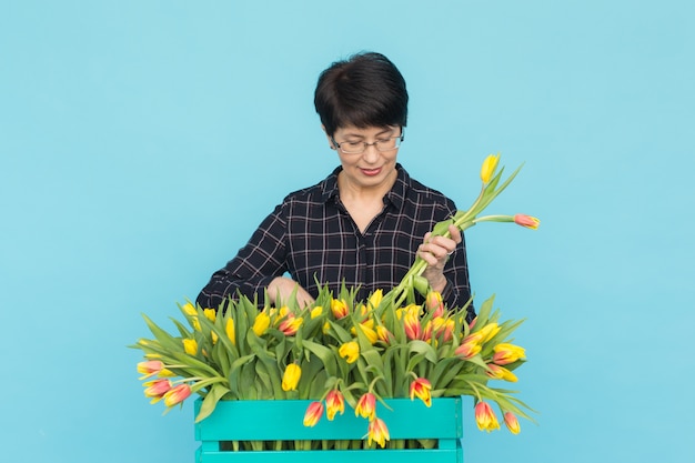 Vrouwelijke bloemist die op middelbare leeftijd blauwe houten doos tulpen op blauw houdt