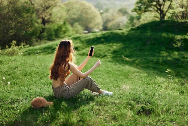 Vrouwelijke blogger zit op het groene gras in een park en maakt foto's van zichzelf op haar telefoon tegen de achtergrond van een zomers landschap Jongeren39s levensstijl en zorg voor het milieu