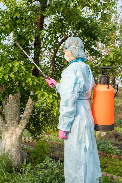 Vrouwelijke boer in een beschermend pak spuit appelbomen tegen schimmelziekte of ongedierte met drukspuit en chemicaliën in de zomerboomgaard.