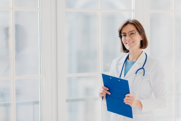 Vrouwelijke cardio-therapeut draagt een ronde bril, een witte medische jas en een fonendoscoop, schrijft met pen in het klembord,