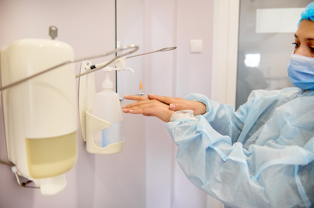 Foto vrouwelijke chirurg die medische beschermende werkkleding en veiligheidsmasker draagt die haar handen vóór chirurgie desinfecteren. steriliteit, voorbereiding op een operatie.