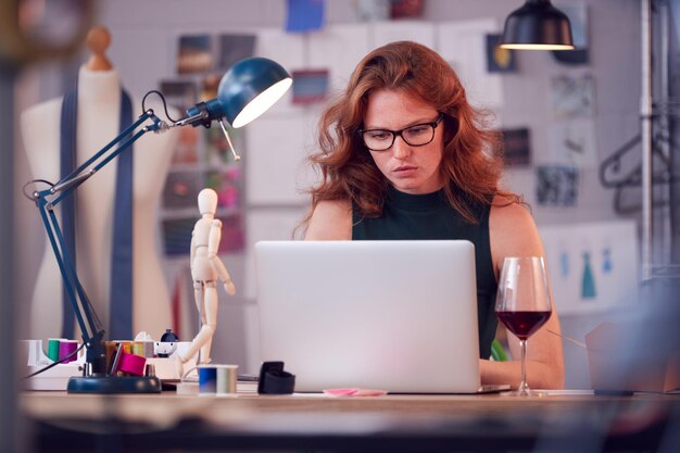 Vrouwelijke eigenaar van modebedrijf met glas wijn laat aan het werk op Laptop In Studio