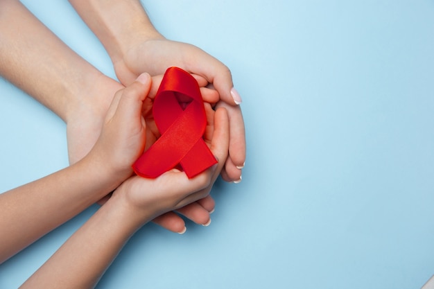 Vrouwelijke en mannelijke handen met rood hiv en aids bewustzijn lint geïsoleerd op blauwe achtergrond. Concept van gezondheidszorg en geneeskunde, wereldwijd ondersteunend, vrijwilligerscampagne tegen ziekte.