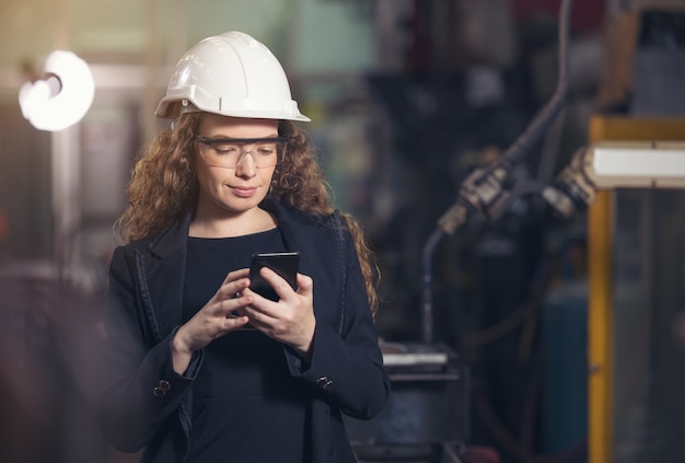 Vrouwelijke fabrieksarbeider die een bouwvakker draagt, gebruikt een mobiele telefoon in een fabriek.