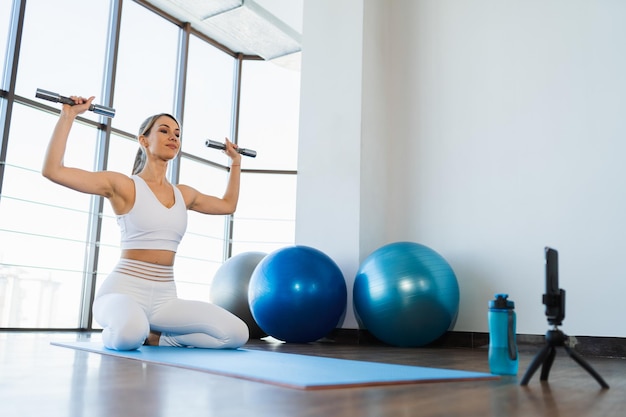 Vrouwelijke fitnesstrainer met halters in witte sportkleding is livestreaming