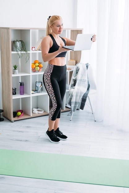 Vrouwelijke fitnesstrainer Thuissport Online oefening gezond lichaam Quarantaine levensstijl Glimlachende atletische vrouw in zwarte sportkleding op zoek naar geopende laptop in handen licht kamer interieur
