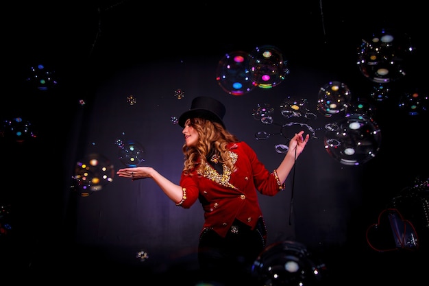 Vrouwelijke goochelaar, een illusionist in theatrale kleding, maakt show met zeepbellen op zwarte achtergrond. Man actrice in toneelkostuum en hoge hoed op haar hoofd. Concept van theatervoorstelling